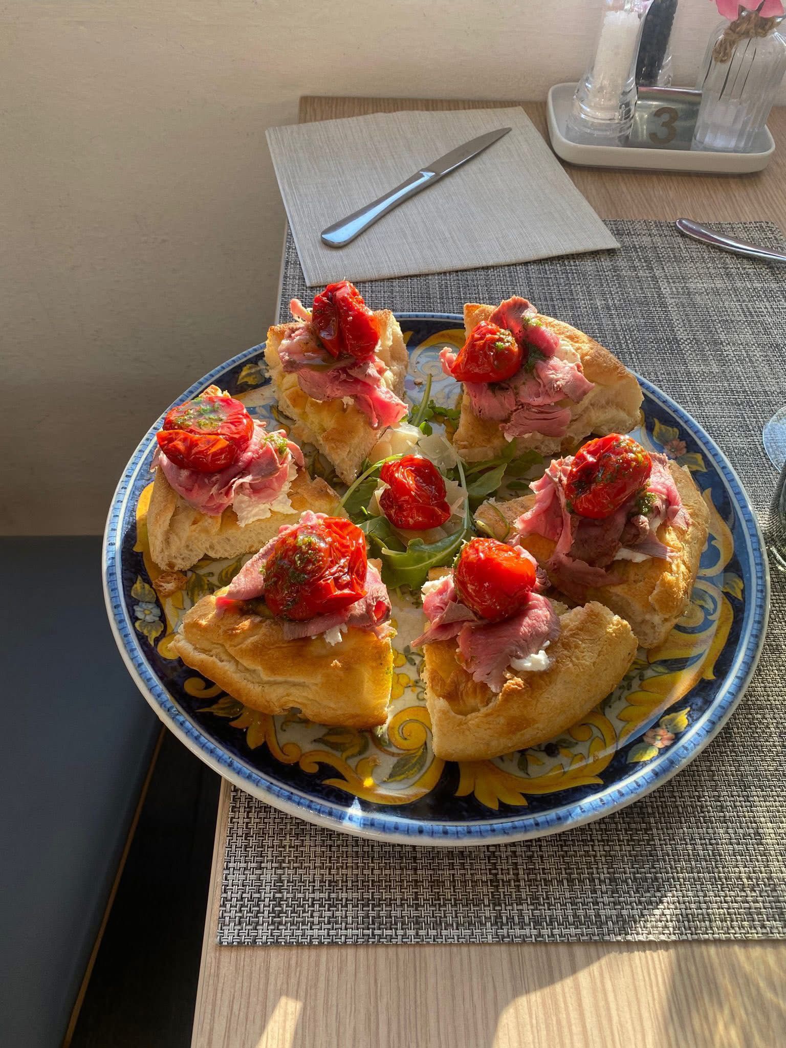 Un piatto decorato con diverse fette di focaccia guarnite con salumi e pomodori secchi, adagiato su un tavolo di legno.