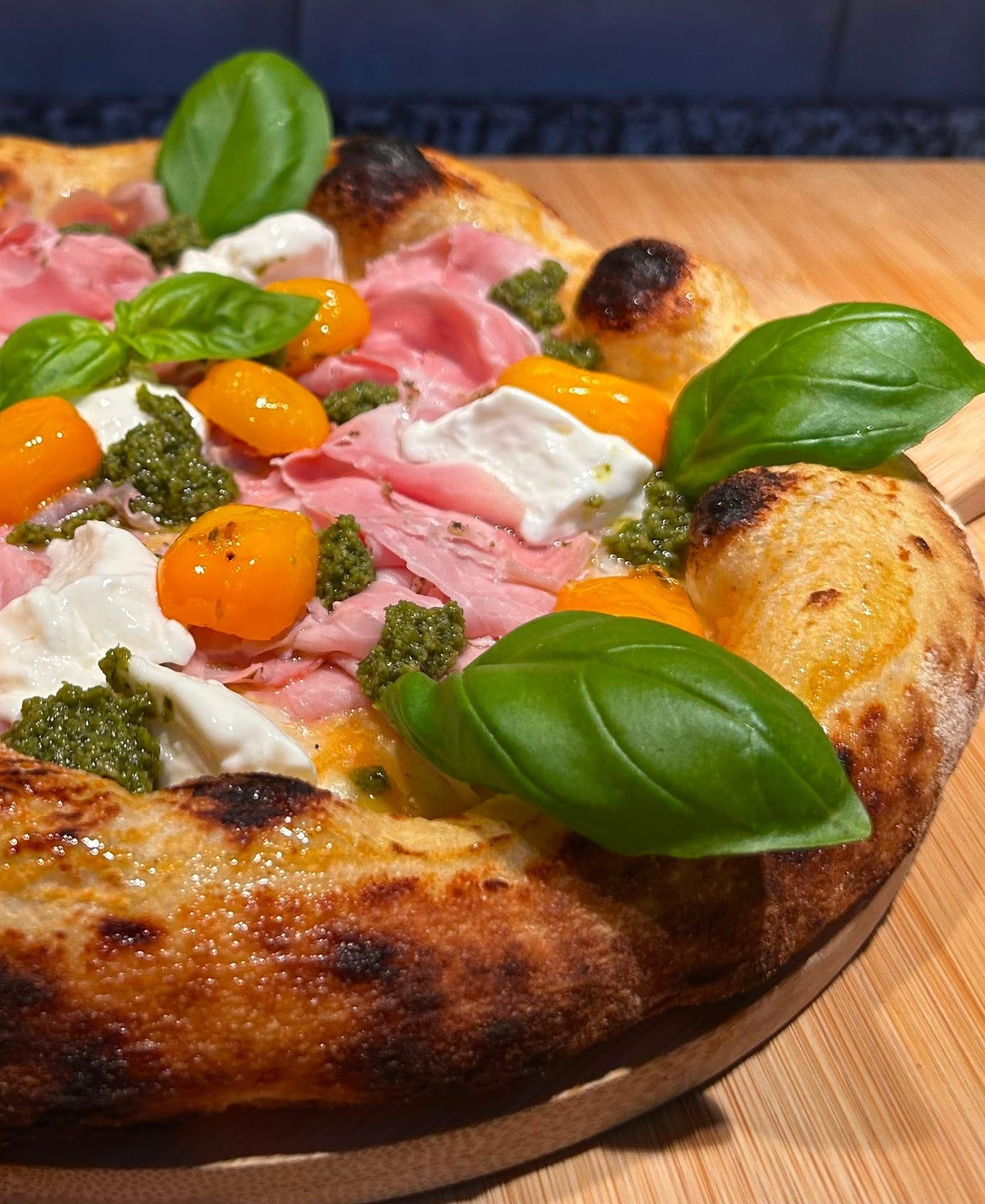 Pizza artigianale condita con prosciutto crudo, pomodori gialli, mozzarella, pesto e foglie di basilico fresco, servita su un tagliere di legno.