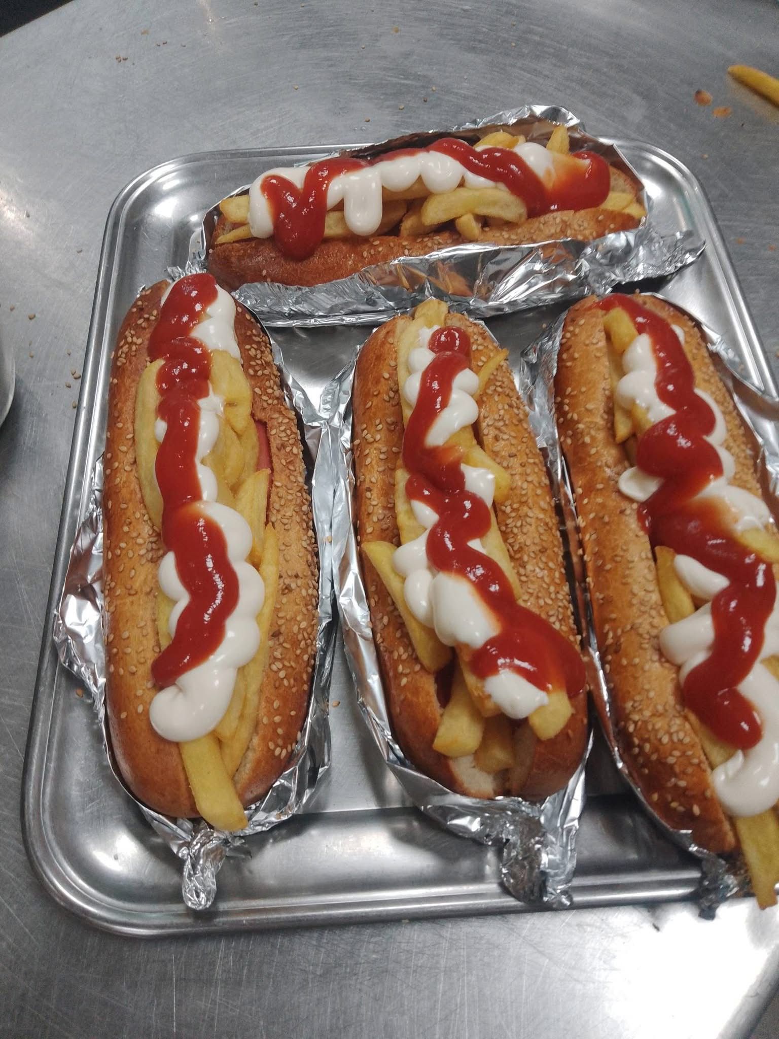 Quattro panini per hot dog ripieni di patatine fritte, conditi con spirali di ketchup e maionese, su un vassoio d'argento.