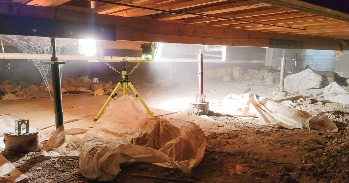Basement crawl space with work lights, dirt floor, insulation, and construction materials under exposed beams