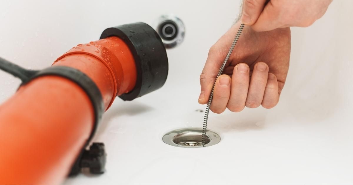 A person uses a thin metal drain snake tool to clear debris from a white bathtub drain.