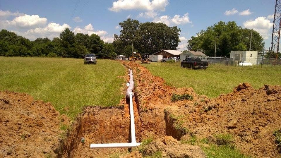 Trenched utility line crossing a grassy field with pipes, dirt piles, and vehicles in the distance
