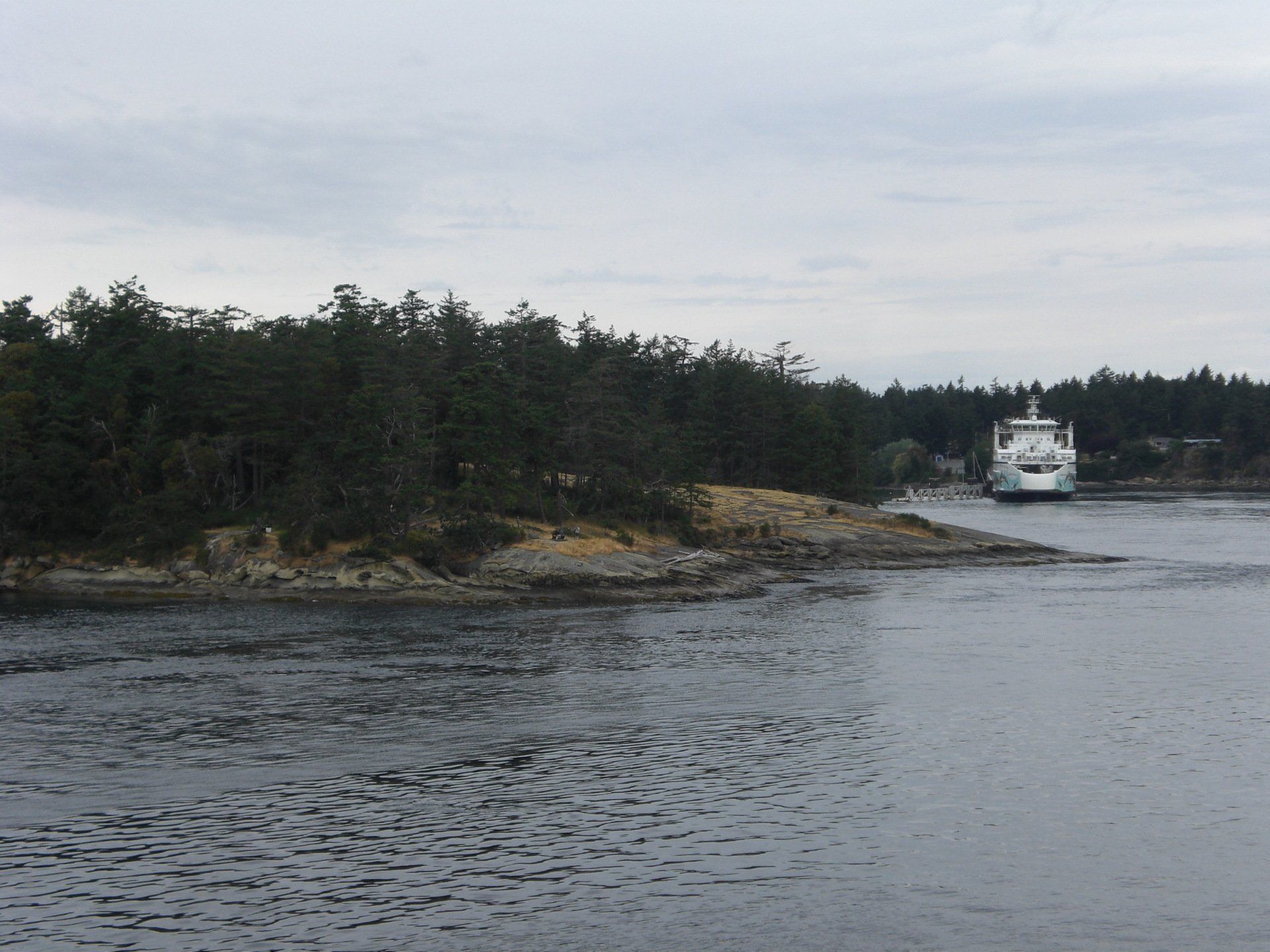 Ferry approaching a rocky shoreline covered in dark green trees under a cloudy sky.