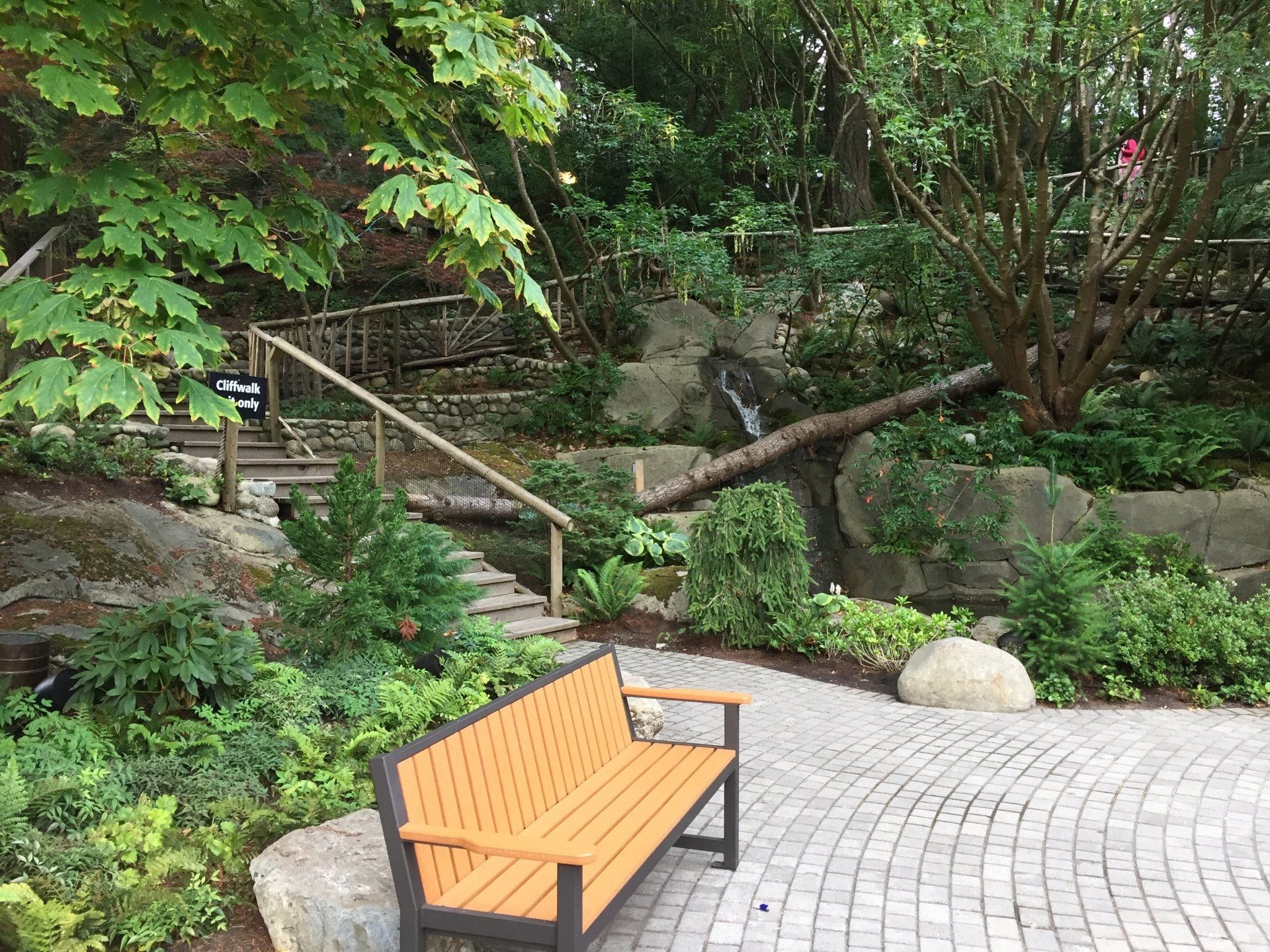 Bench in a lush garden setting with stone steps, wooden railing, and a small waterfall.