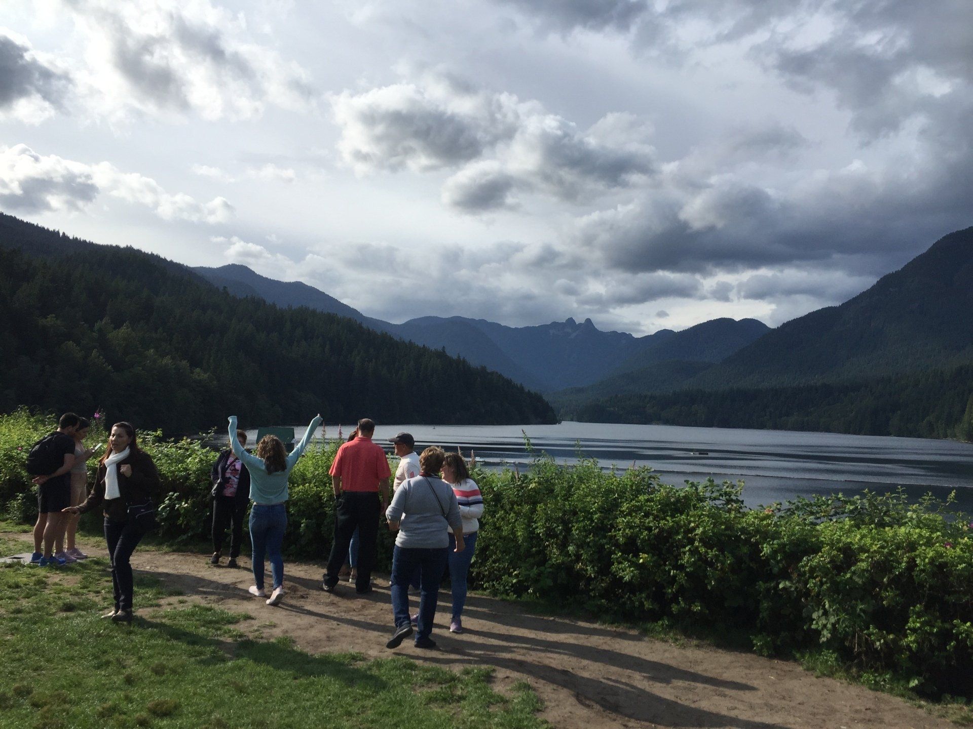 Group of people overlooking a lake and mountain range under a cloudy sky.