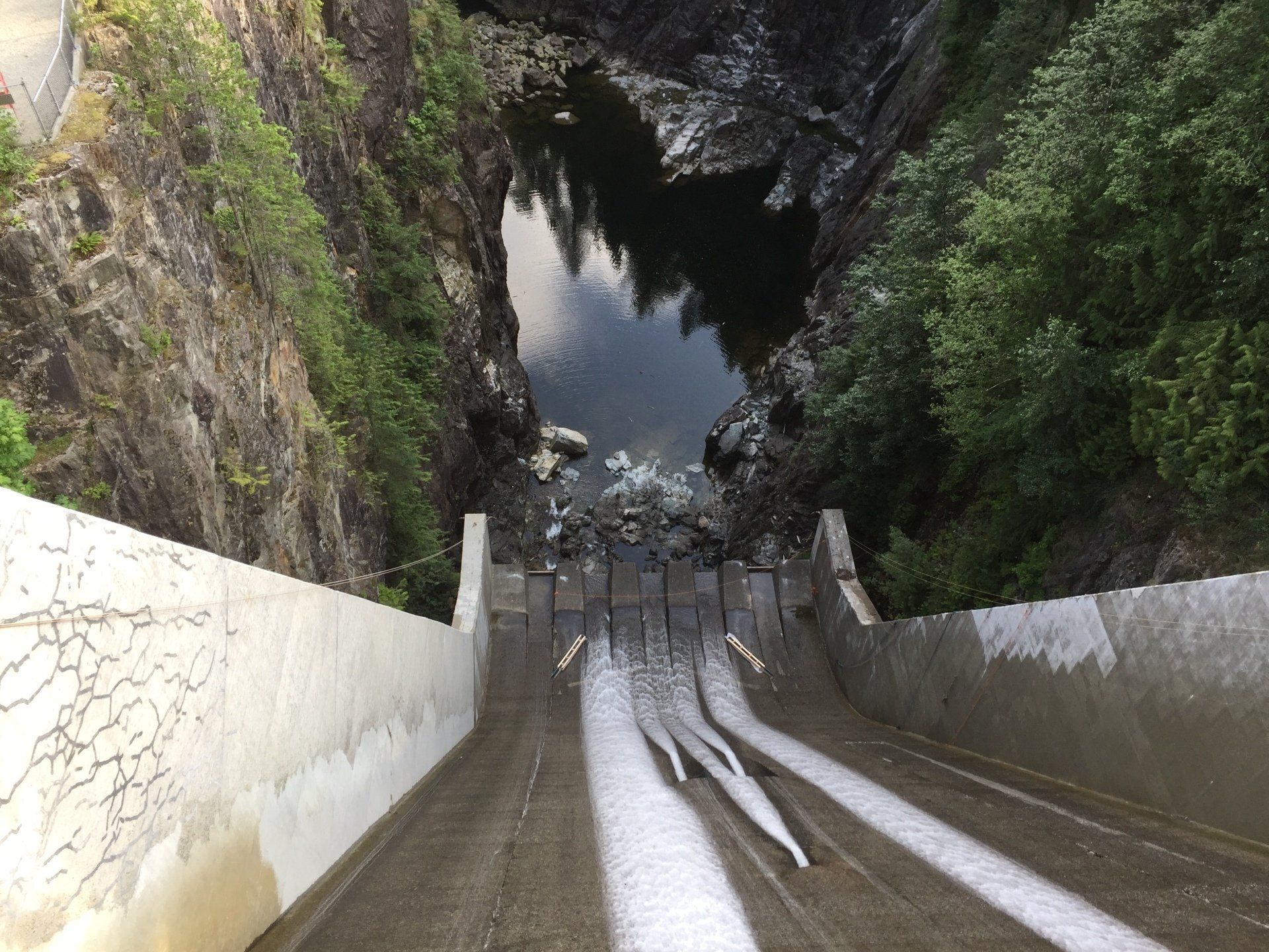 A concrete dam with water flowing down a long slope into a dark pool surrounded by forest.