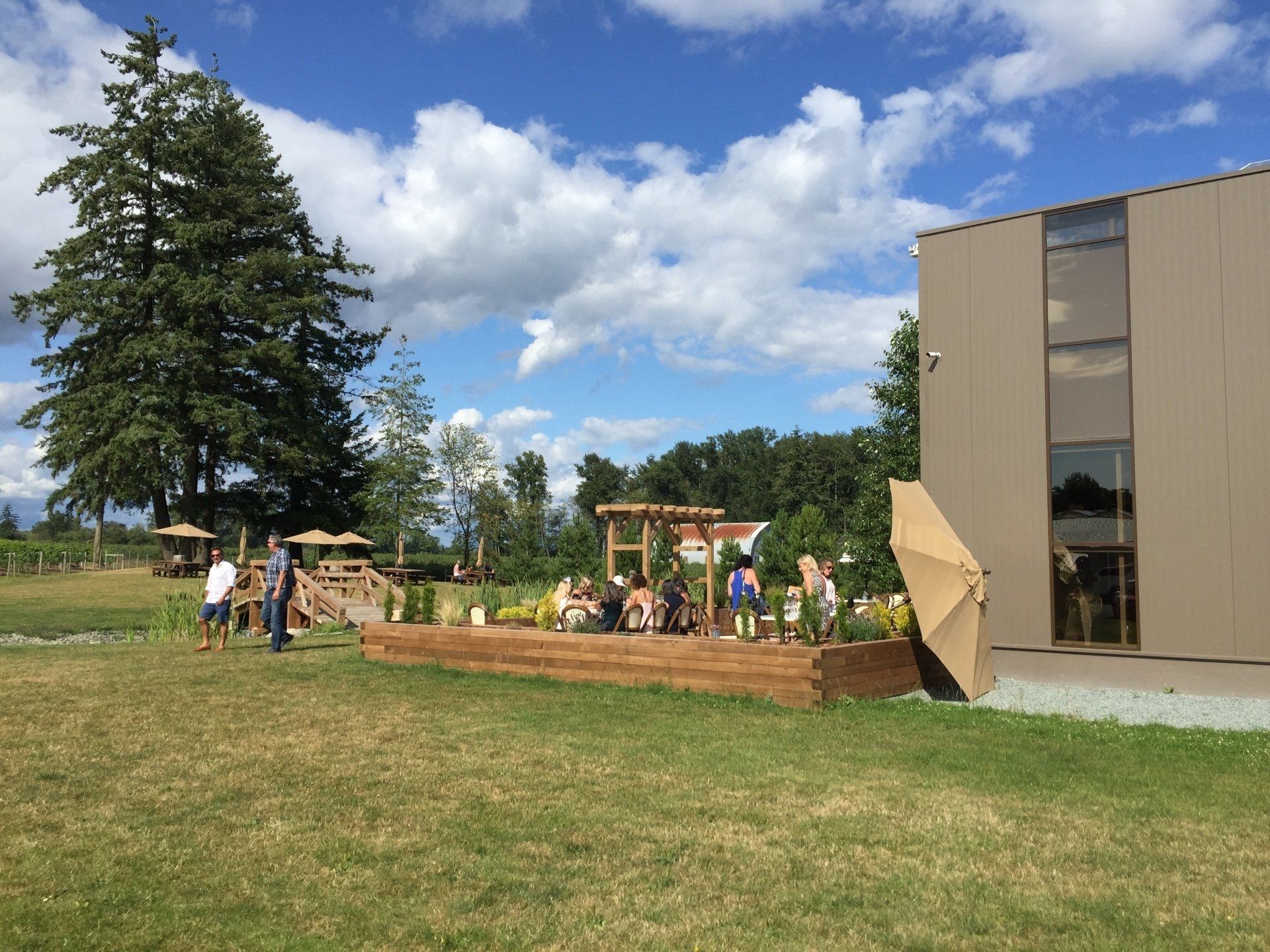 People on a wooden patio with a building on the right and trees and sky in the background.
