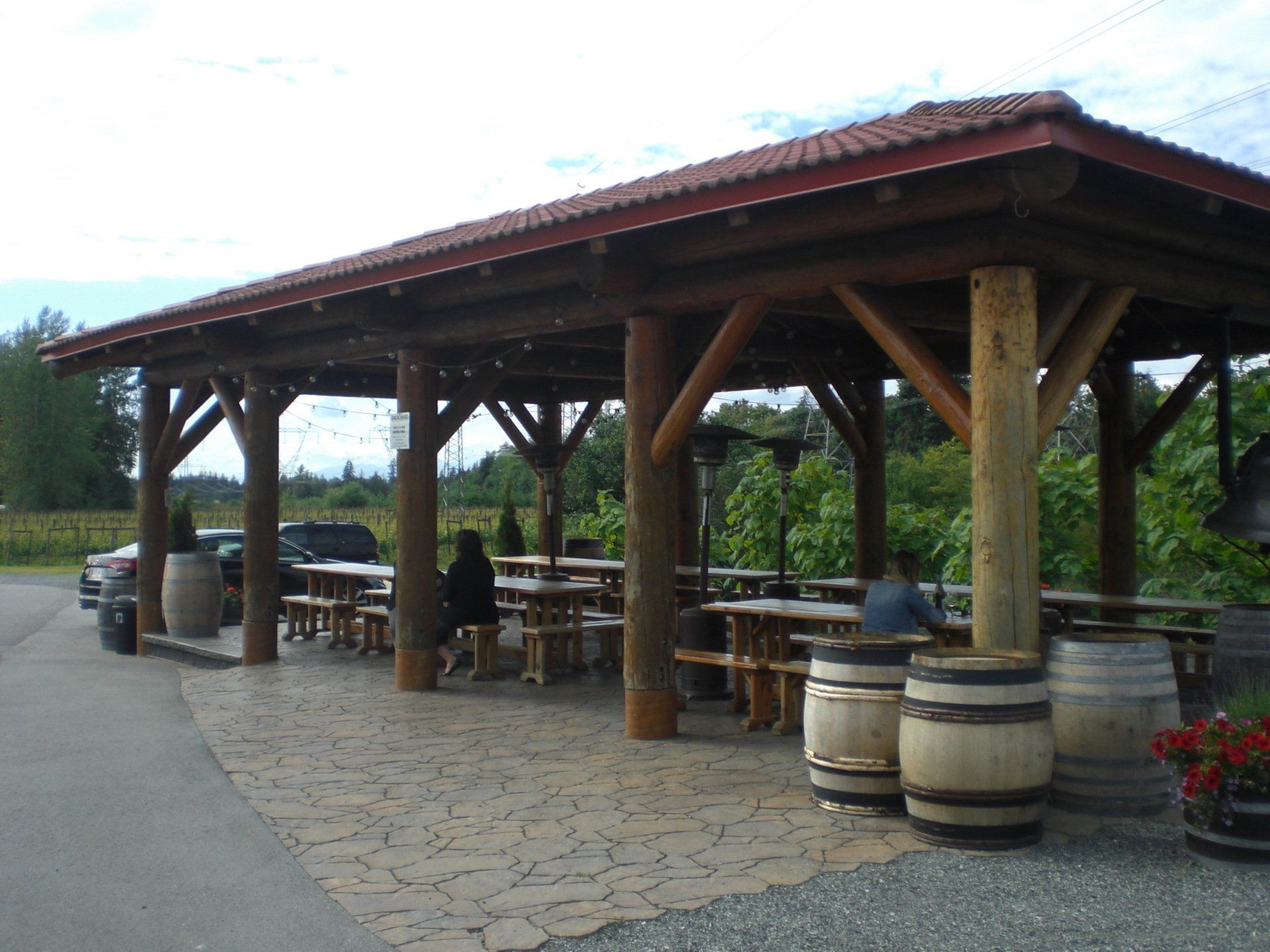 Outdoor wooden shelter with picnic tables and wine barrels, possibly at a vineyard.