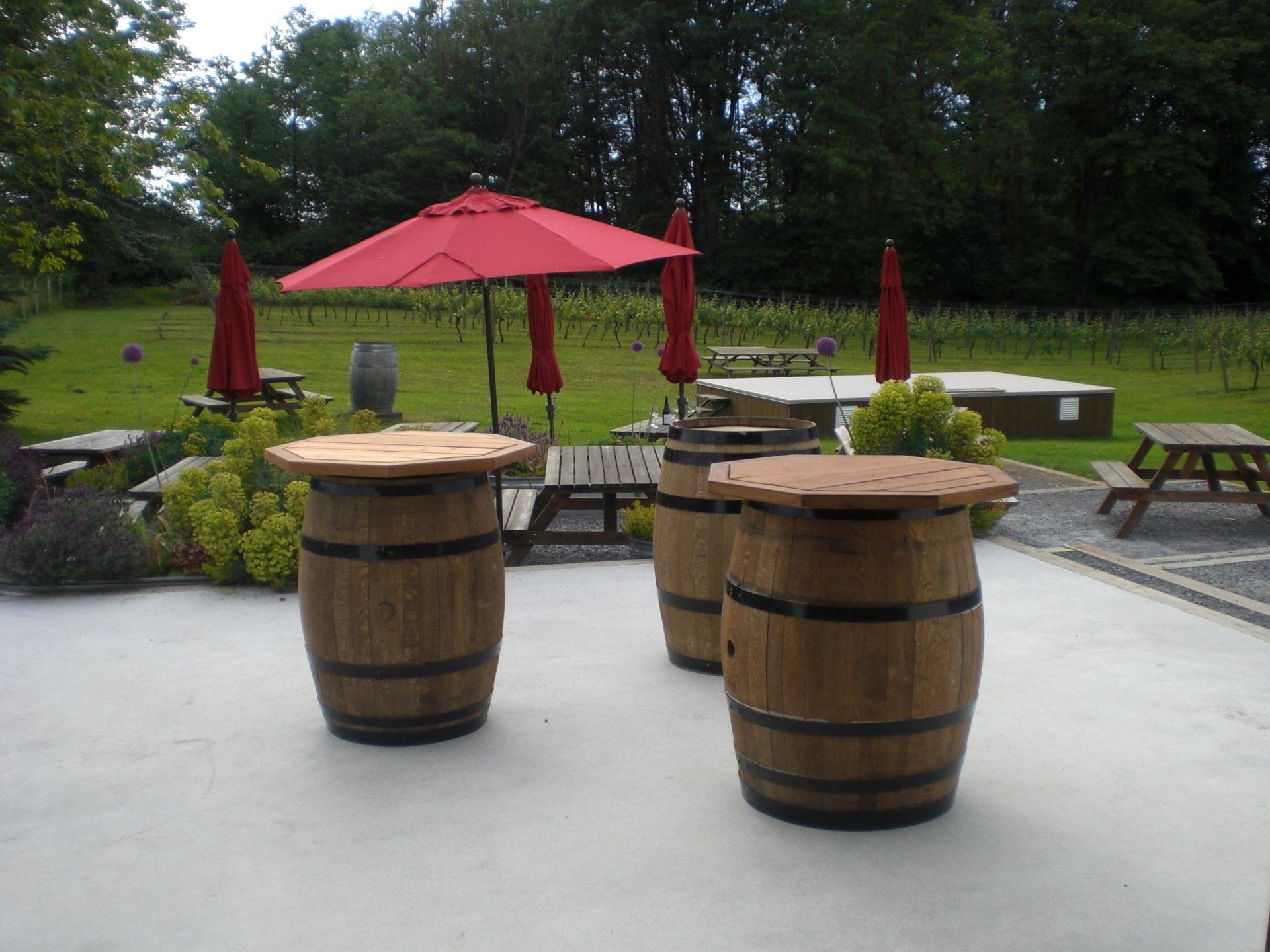Three wooden barrel tables with red umbrellas on a patio overlooking a vineyard.
