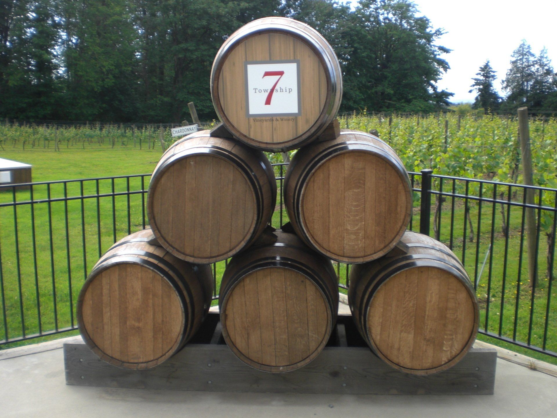 A stack of six wooden barrels with a seventh barrel on top with the number seven in front of a vineyard.