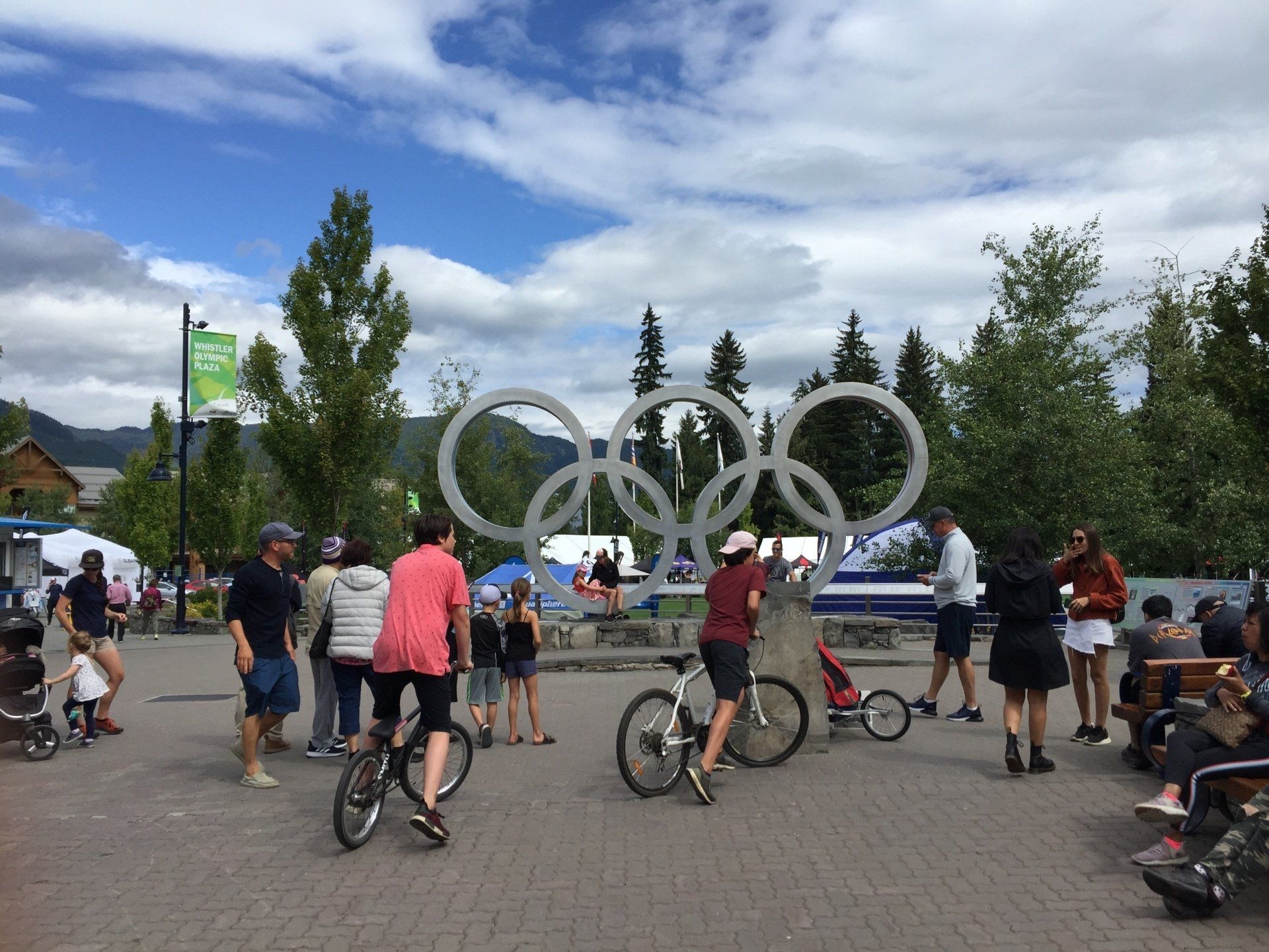 People gather near Olympic rings in Whistler, BC. Sunny day with bicycles, trees, and shops.