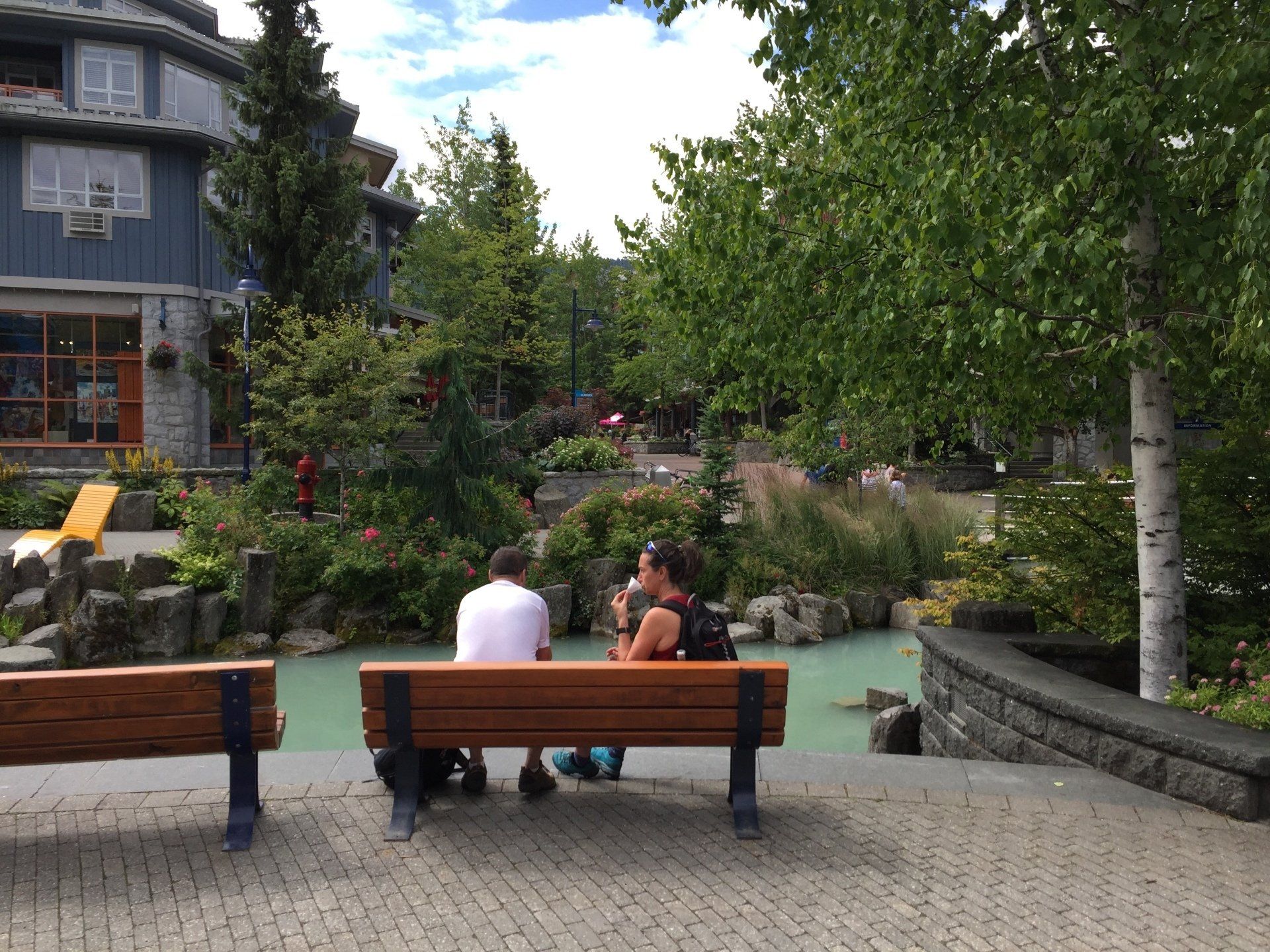 Two people on a bench by a light blue water feature, trees and buildings in the background.