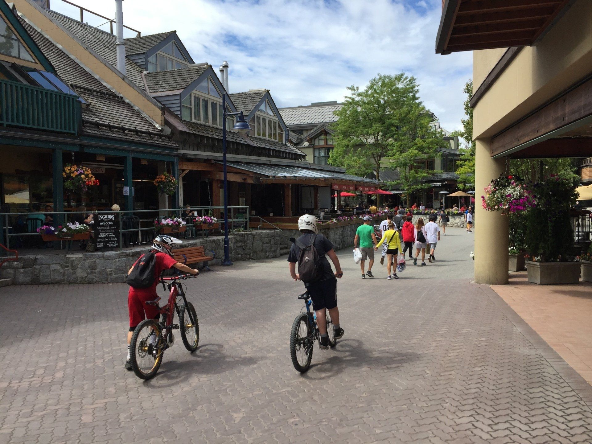 Two cyclists ride on a cobblestone path lined with shops and people in a village square.