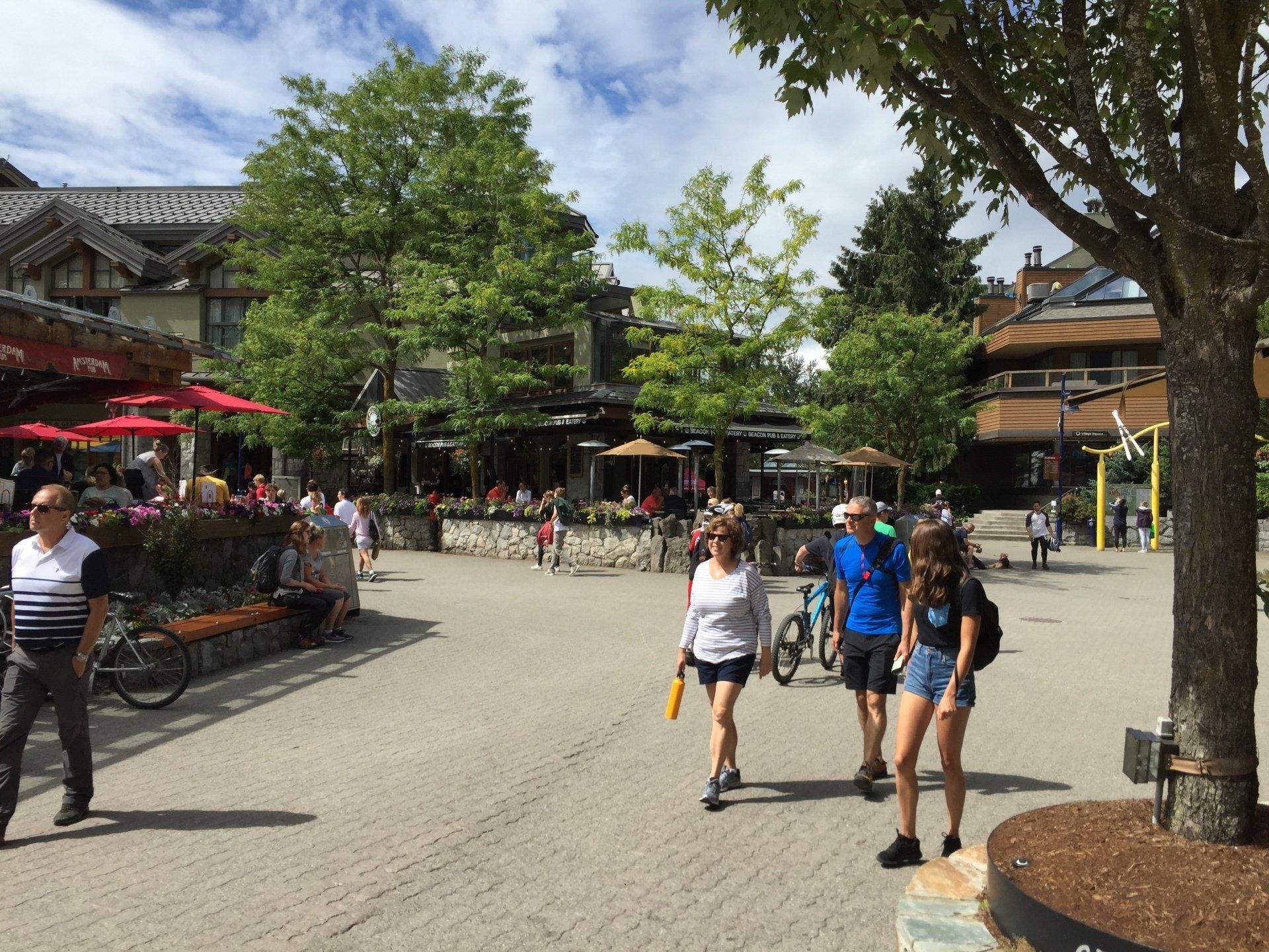 People walking in a sunny plaza lined with shops and restaurants. Trees and blue sky in the background.