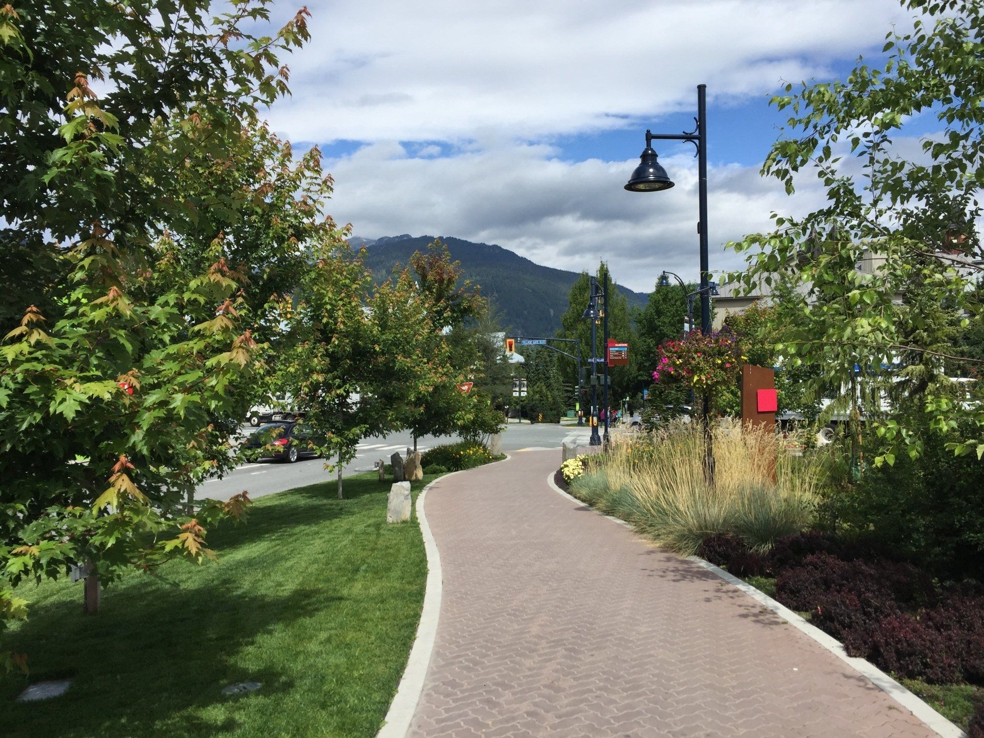 A paved walkway winds through green trees, leading toward a street and mountain on a cloudy day.