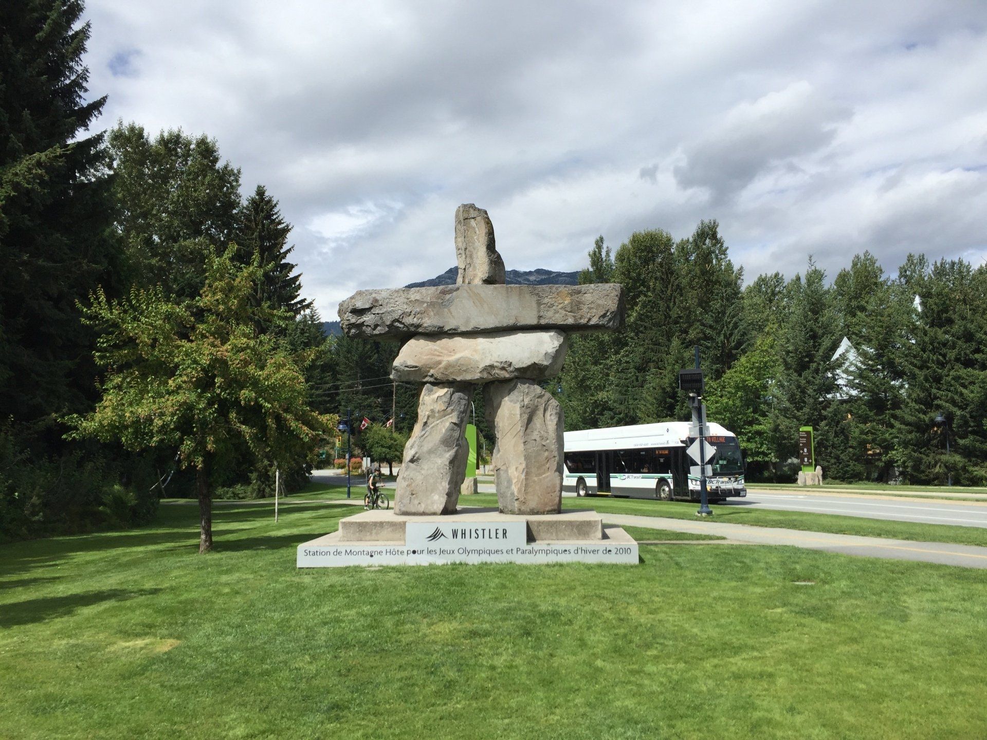 Stone Inukshuk sculpture on a grassy area with trees and a bus in the background.
