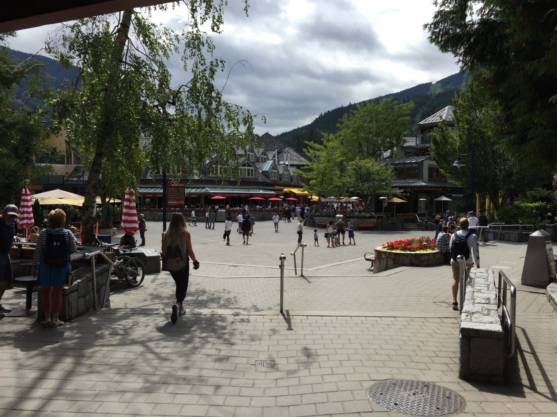 A pedestrian plaza in a mountain village with people walking, trees, and buildings under a cloudy sky.