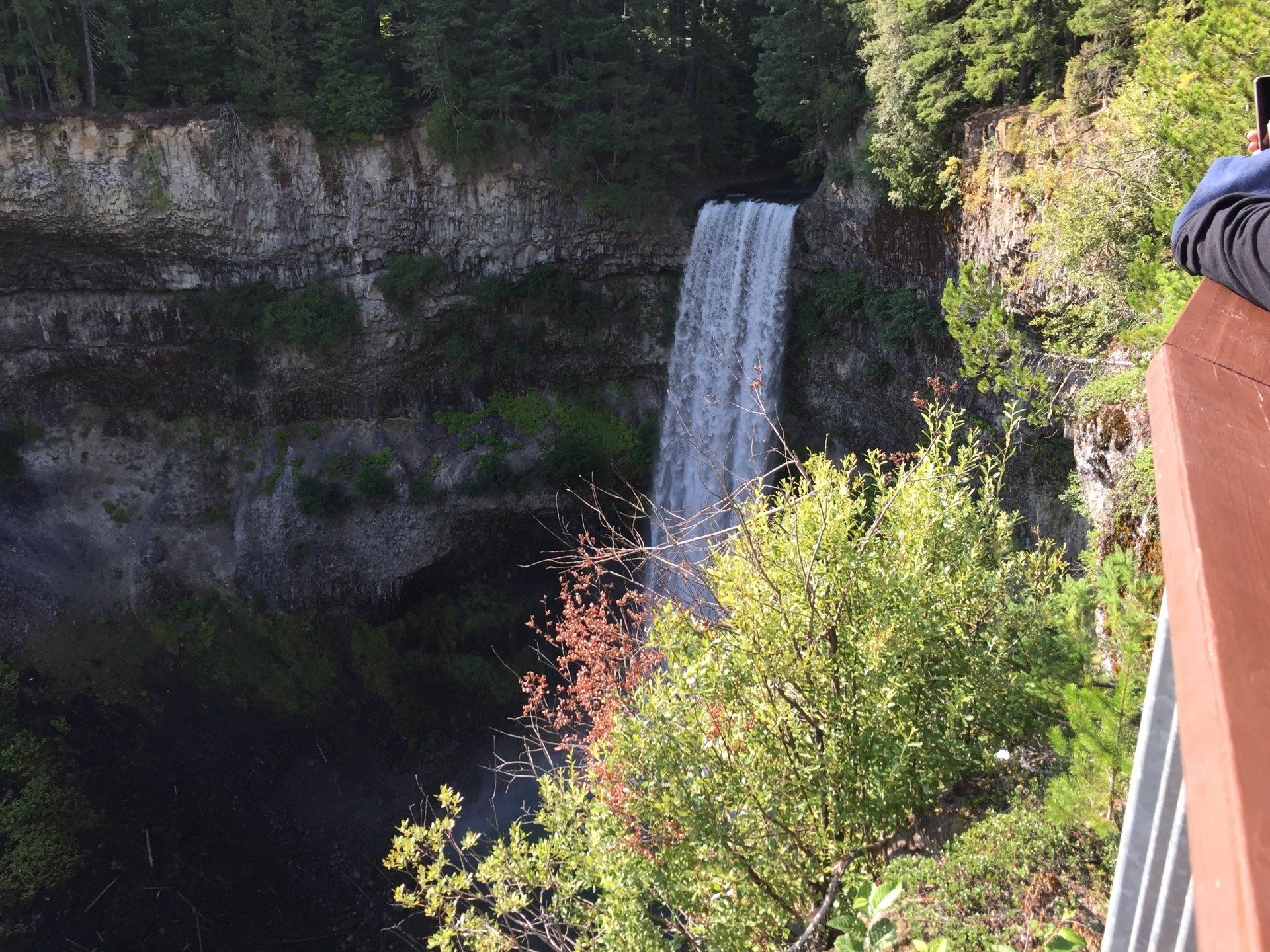Waterfall cascading down a cliff into a dark pool, viewed from a wooden railing.