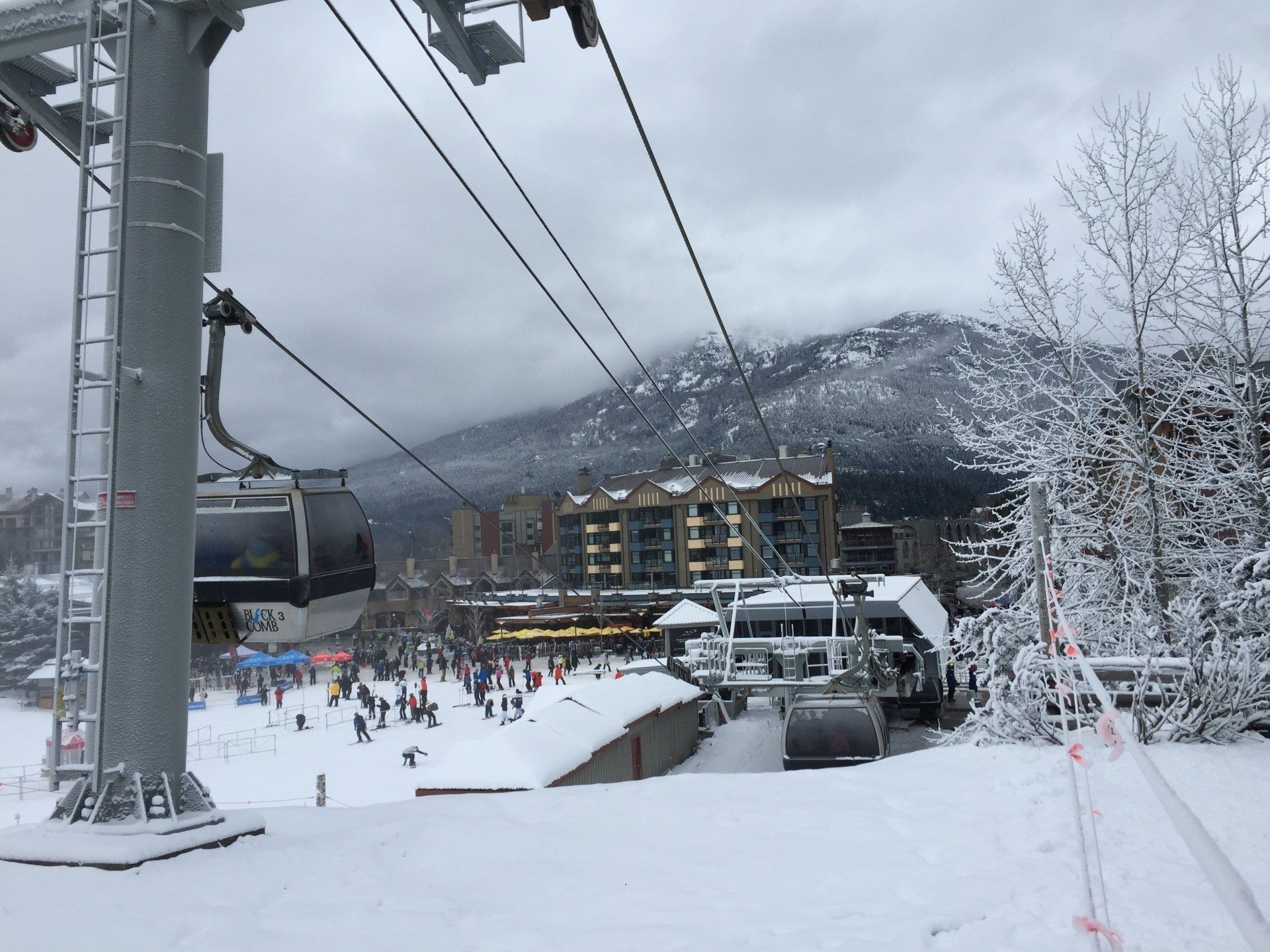 Ski lift above a snow-covered ski resort, with buildings and people in the distance. Overcast sky.