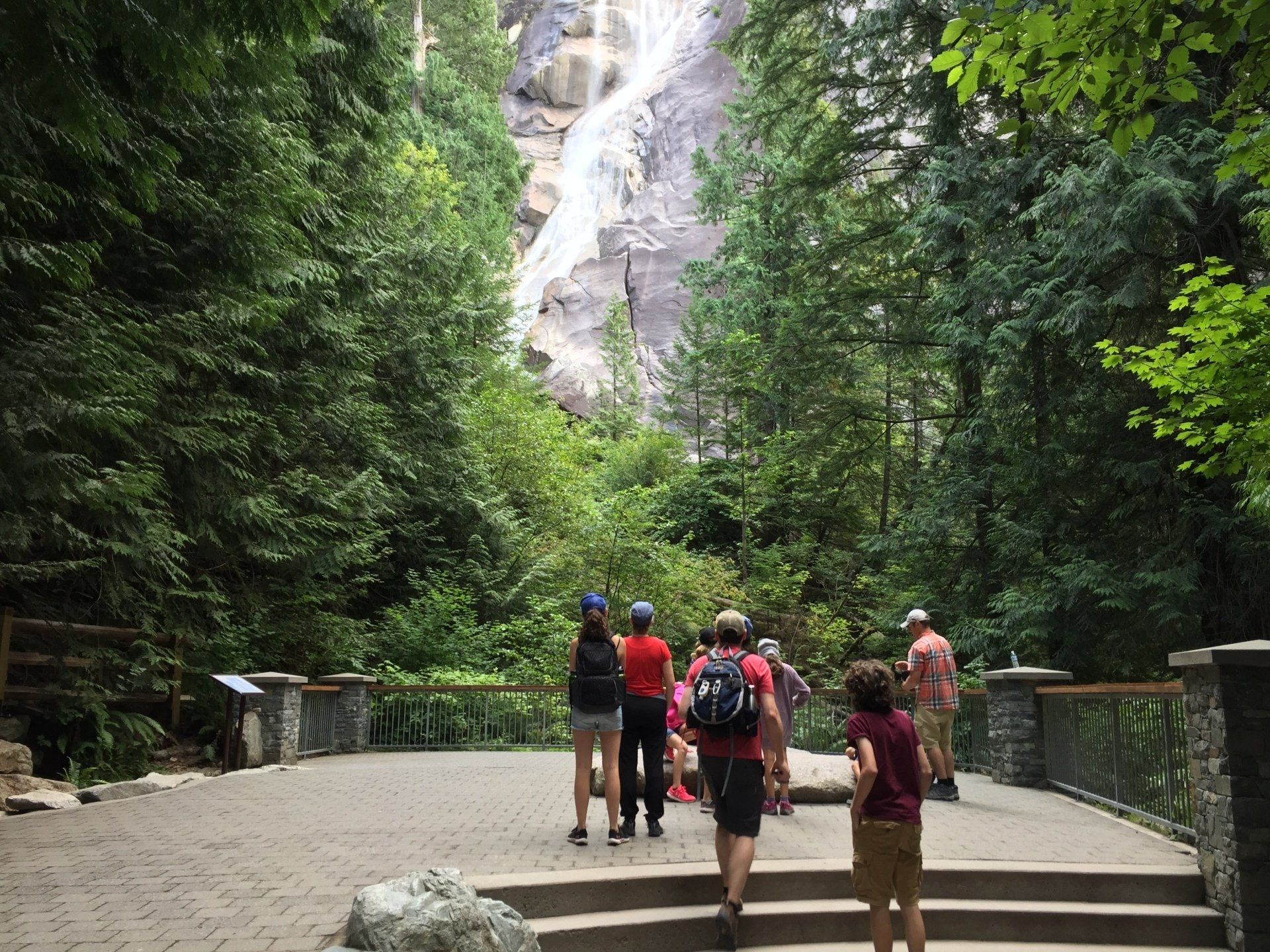 People viewing a tall waterfall from a stone pathway in a lush green forest.