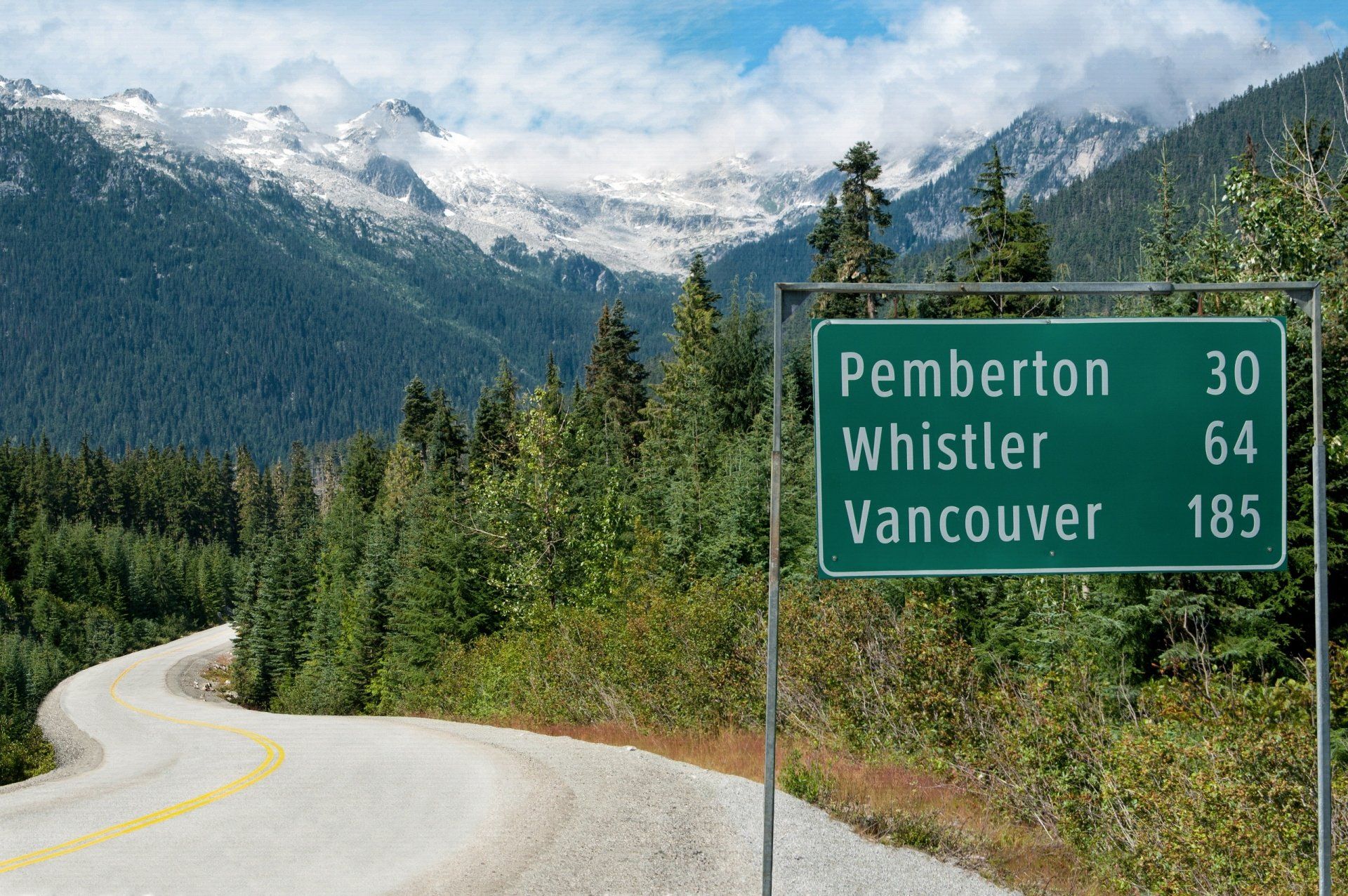 Road sign indicating distances to Pemberton, Whistler, and Vancouver, with a mountain backdrop.