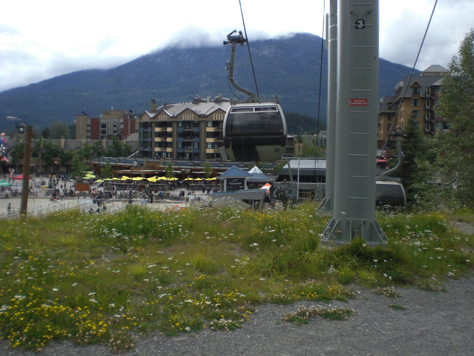 Cable car ascending toward mountain peak, over a village square with people and buildings.