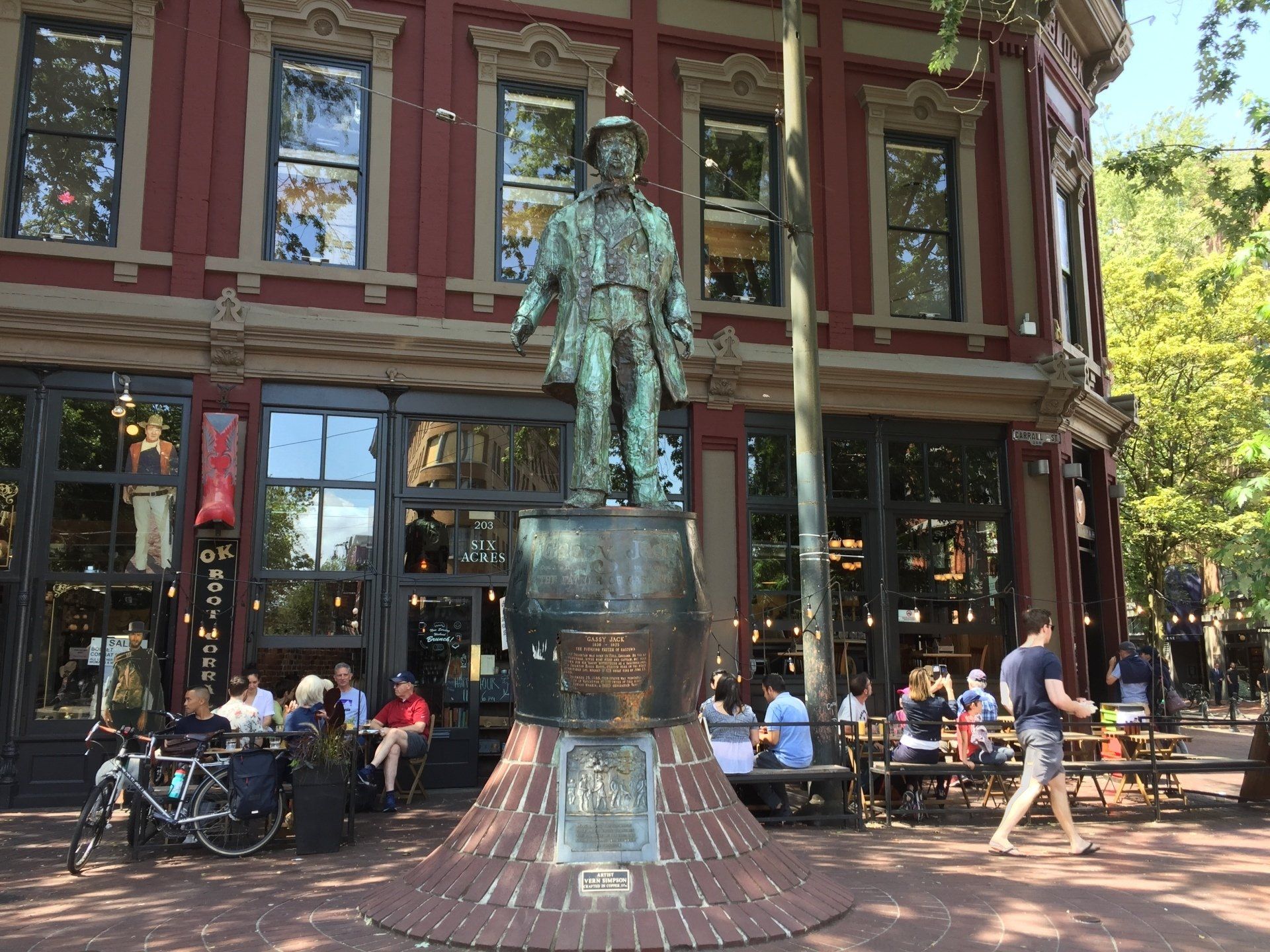 Statue in front of a red building; people seated at outdoor tables, green trees, sunny day.