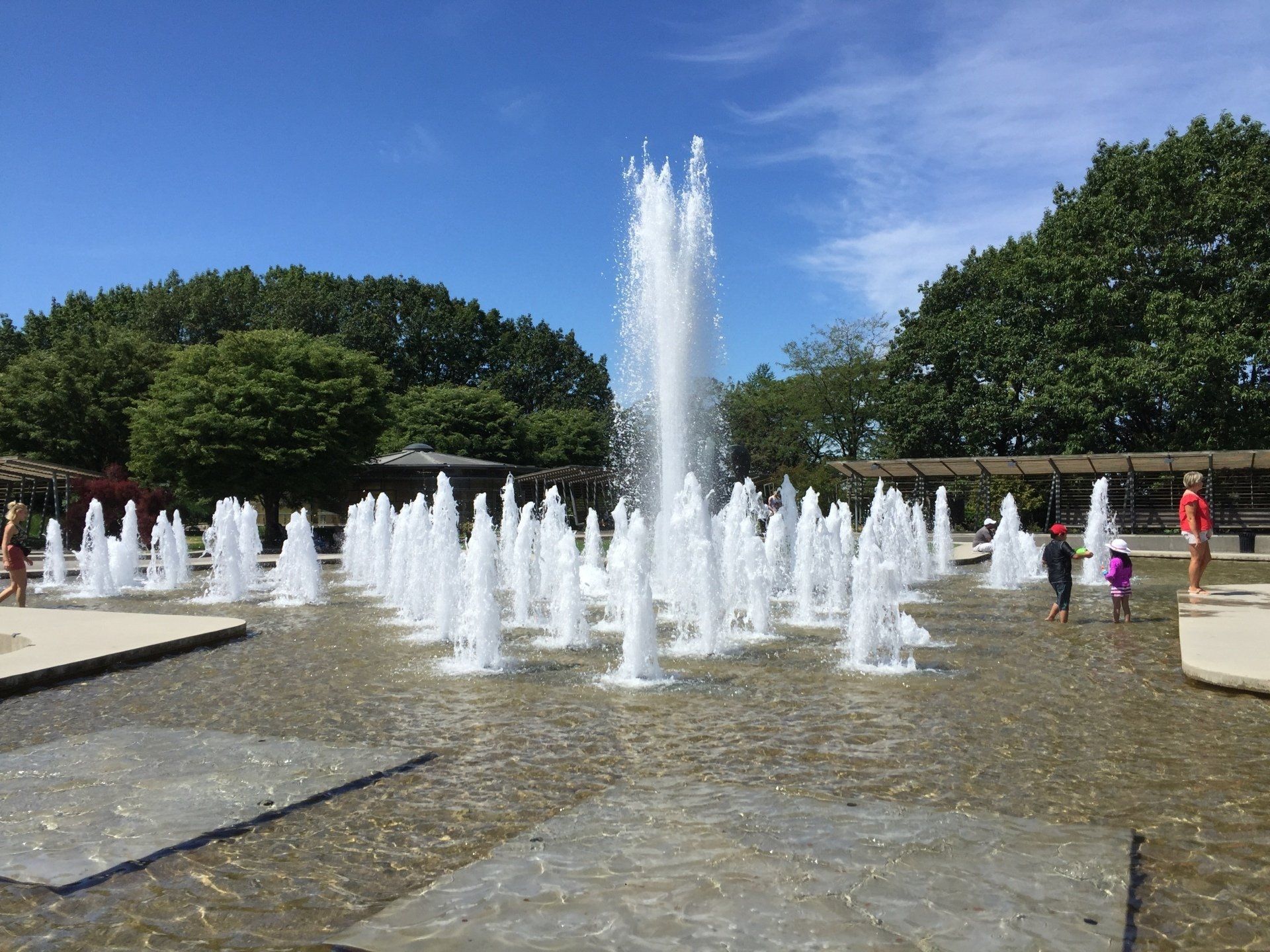 Fountain spraying water into the air in a park on a sunny day. Children play nearby.