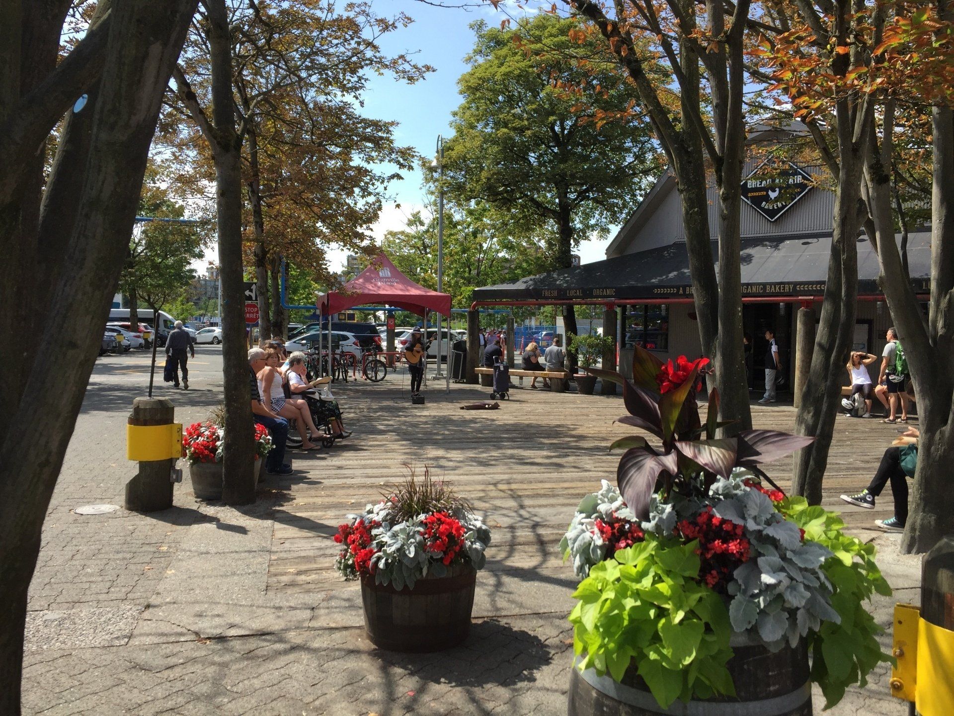 Outdoor scene: people sitting and walking. Red tent, flower barrels, and storefront.