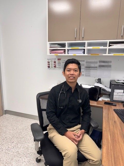 A man wearing a pink shirt and a stethoscope around his neck is sitting in front of a computer.