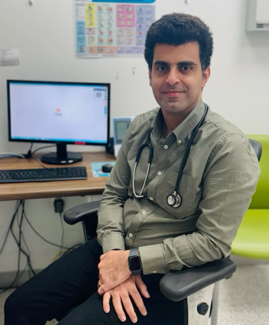 A man wearing a pink shirt and a stethoscope around his neck is sitting in front of a computer.