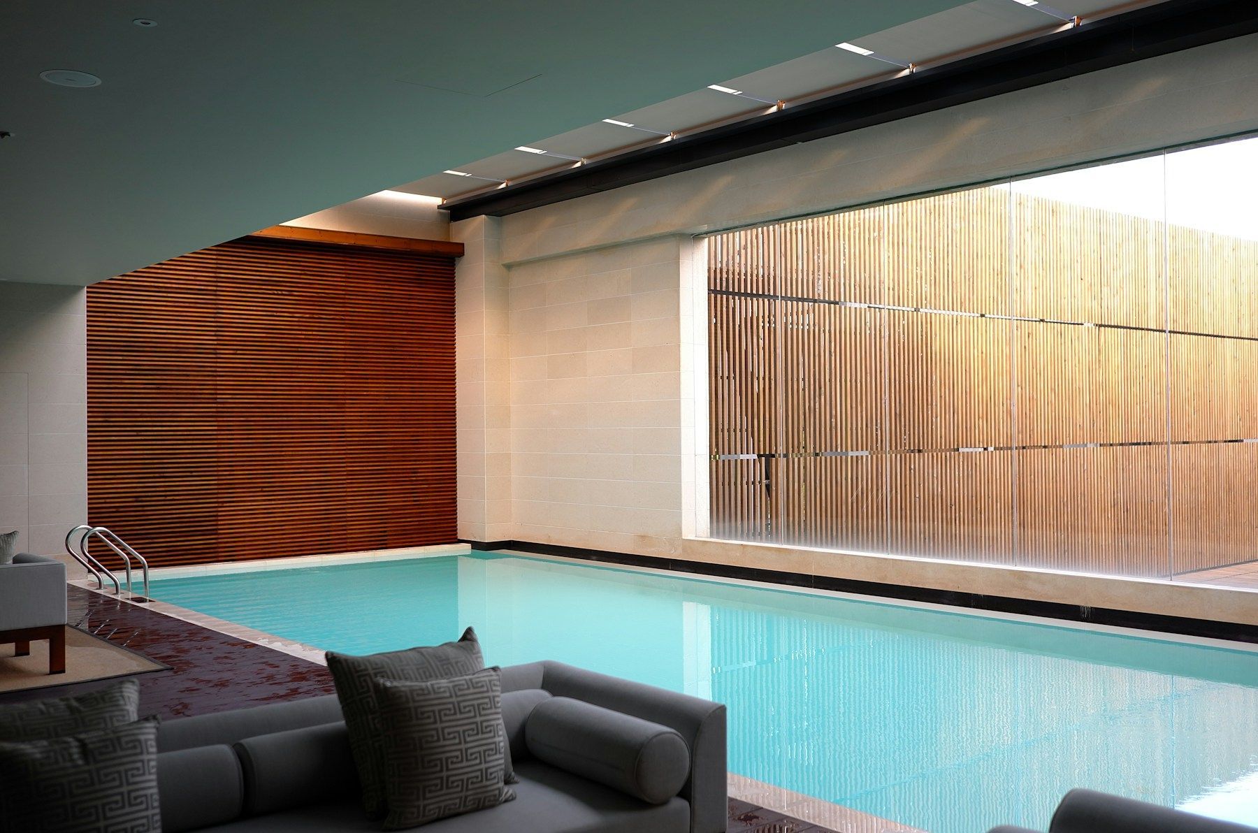 Modern indoor pool with turquoise water, lounge chairs, and wood-accent wall panels