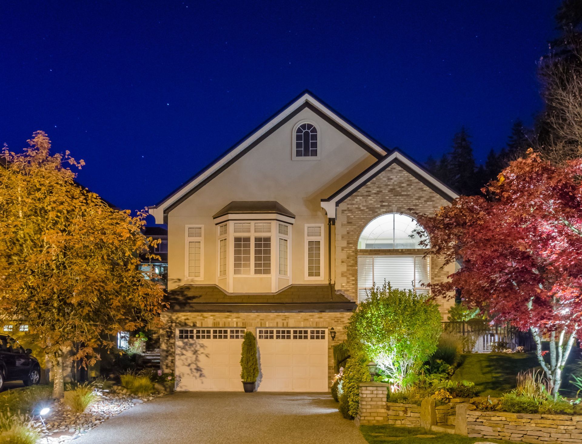 Two-story house with lit windows and garage at night, framed by colorful autumn trees and a deep blue sky.