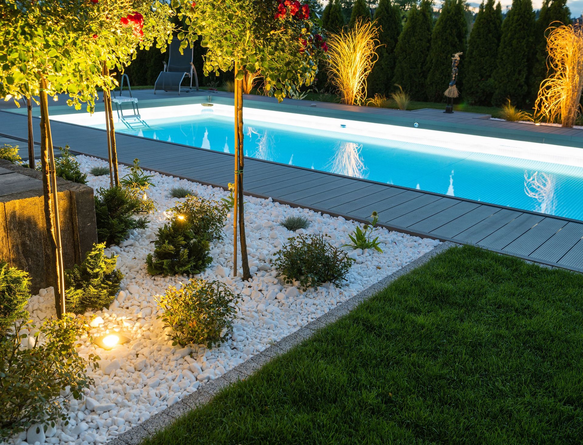 Lit backyard pool beside landscaped gravel garden and green lawn at dusk