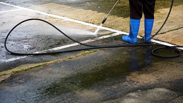 Person in blue boots pressure washing a wet, outdoor surface near white lines.