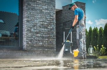 Man power washing a concrete surface next to a brick building.