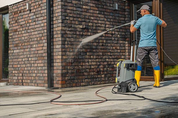Person pressure washing a brick wall outside a house.