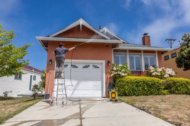 Man on ladder pressure washing a two-story orange stucco house with white garage door on a sunny day.