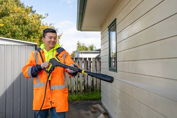 Man power washing a house with a pressure washer; he wears a safety vest, outside.