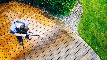 Man pressure washing a wooden deck, with contrast between cleaned and uncleaned sections.