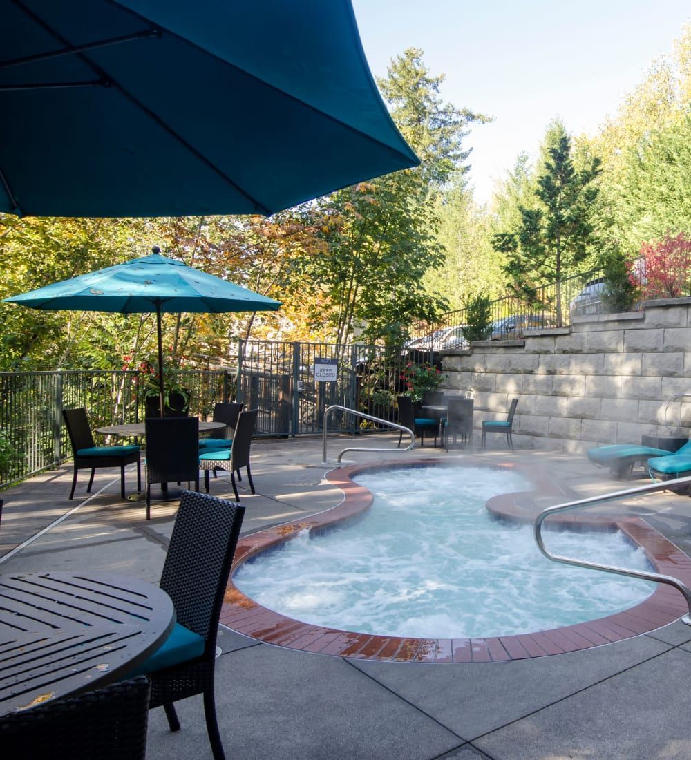 Hot tub with bubbling water, patio furniture, and teal umbrella in outdoor setting with trees.