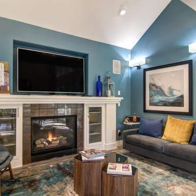 Living room with fireplace, TV, and blue walls. Gray sofa, wooden coffee tables, and a colorful rug complete the space.