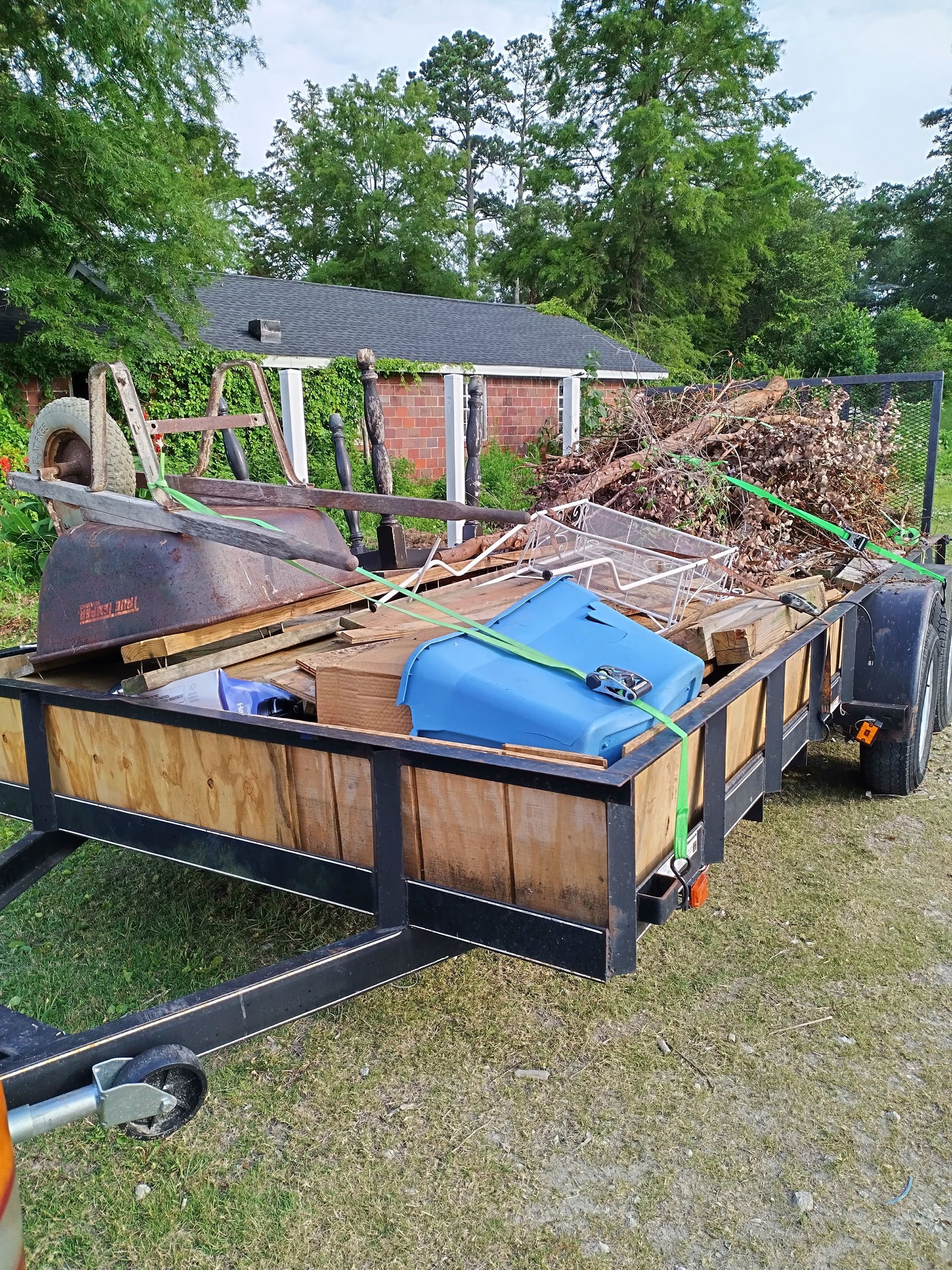 A trailer filled with wood and a blue tank is parked in a grassy area.