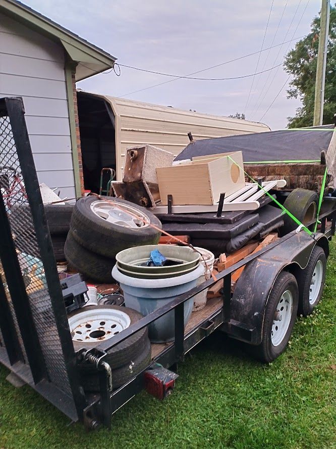 A trailer filled with junk is parked in the grass in front of a house.