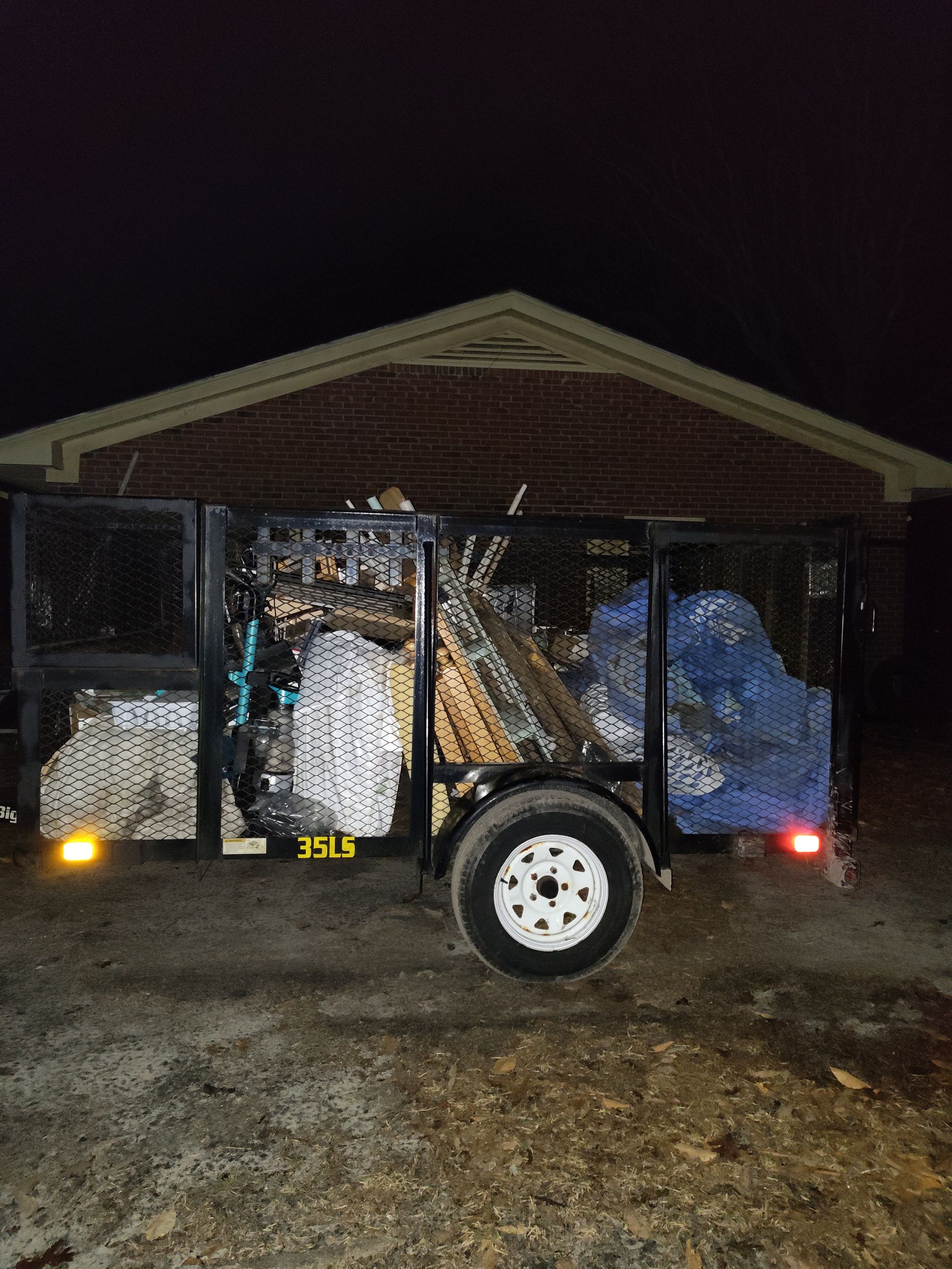 A trailer filled with trash is parked in front of a brick house at night.