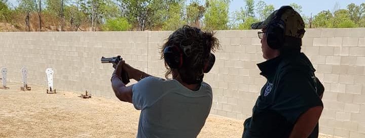 Person firing a handgun at a shooting range. Another person looks on. Steel targets in the distance.