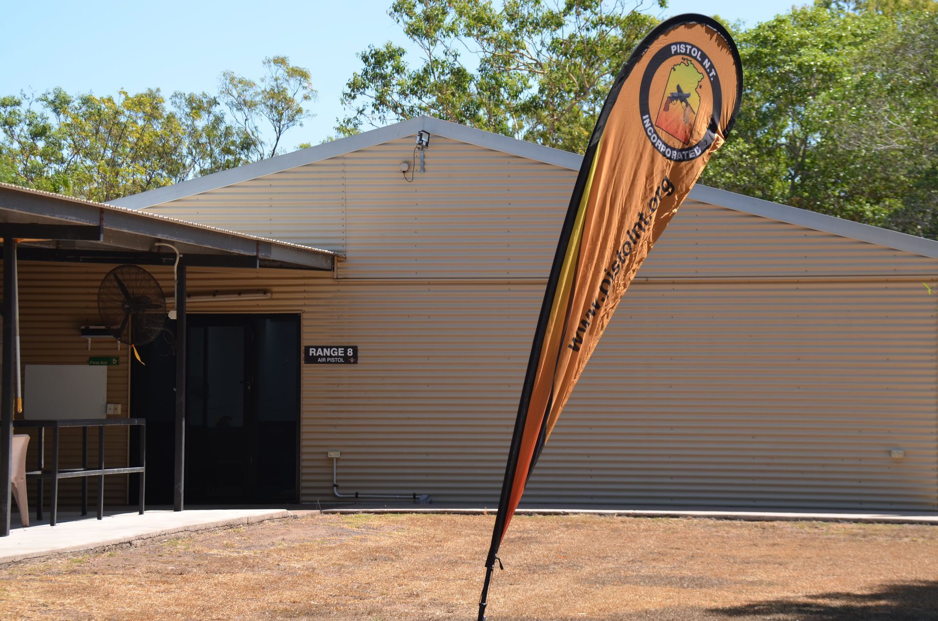 Building with a teardrop flag in front. Brown corrugated metal, trees in background, sunny day.