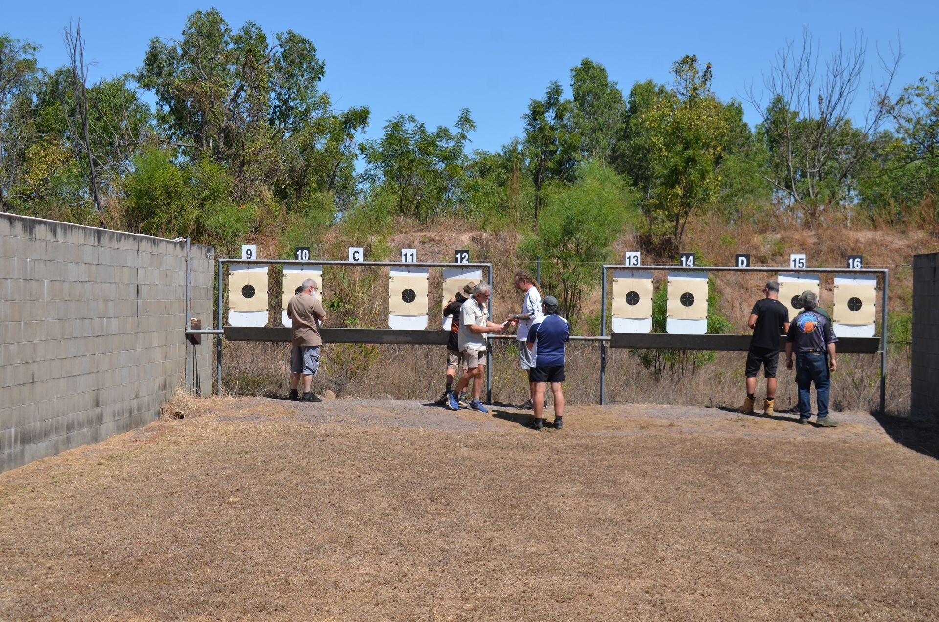 People at a shooting range, firing at targets. Gray concrete walls, brown ground, and trees in the background.