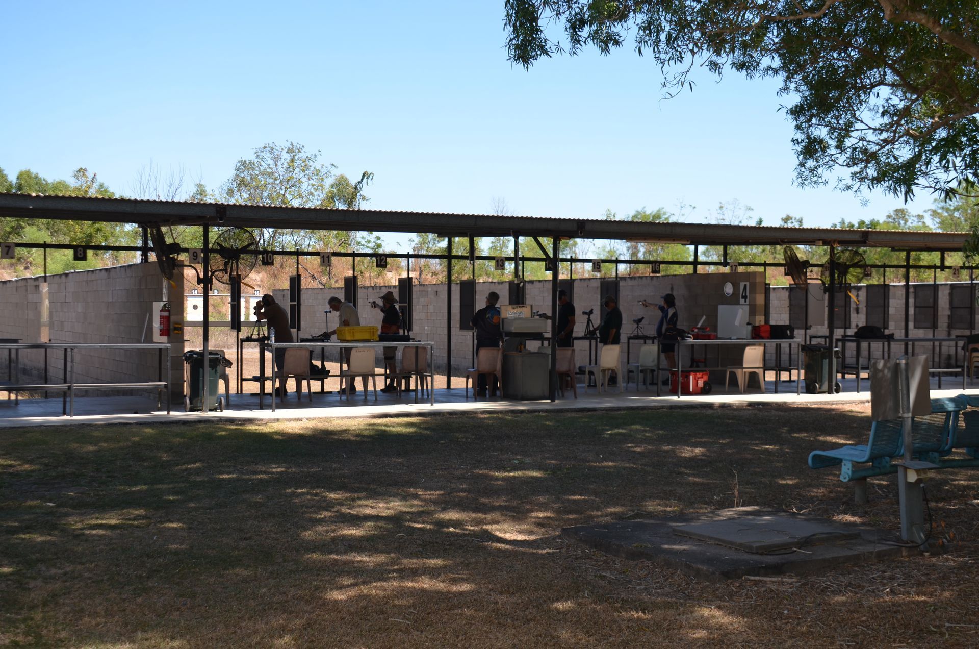 Shooting range with people firing guns under a covered structure on a sunny day.