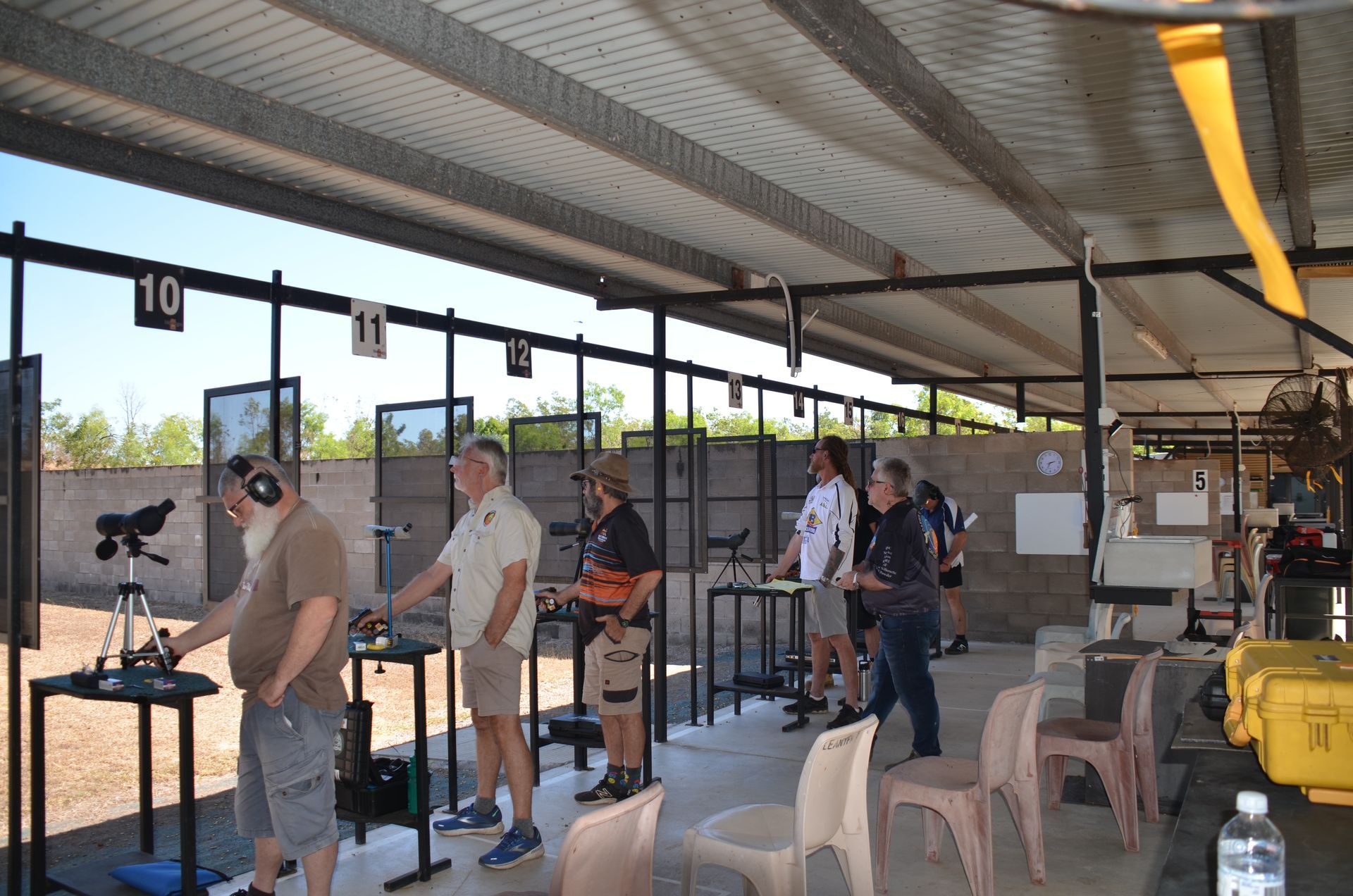 People shooting at a gun range, various ages, under a metal roof, shooting stations marked 1 to 10.